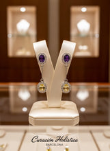 Pair of earrings with purple gemstones on a white stand, with 'Curacion Holistica Barcelona' branding.
