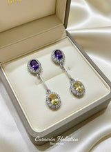 Pair of earrings with purple and yellow gemstones in a jewelry box, branded 'Curación Holástica'.