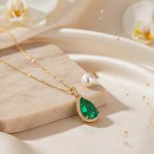 Gold necklace with a green teardrop pendant on a beige surface with flowers in the background