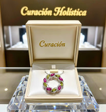 Jewelry box with a ring featuring green and pink gemstones on a reflective surface with 'Curación Holística' branding in the background.